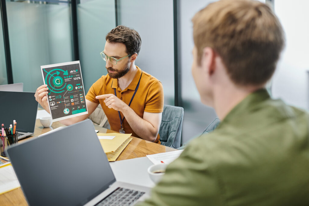 businessman pointing at infographics near laptops and colleague in modern office